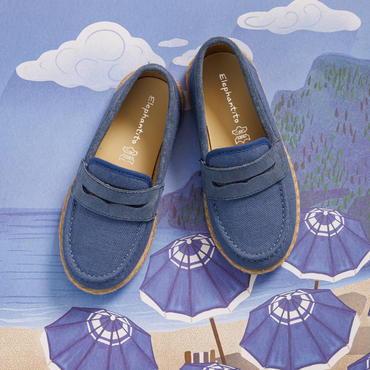 Elephantito - Indigo blue toddler loafer espadrilles with stitched detailing and tan rubber sole on illustrated beach background.