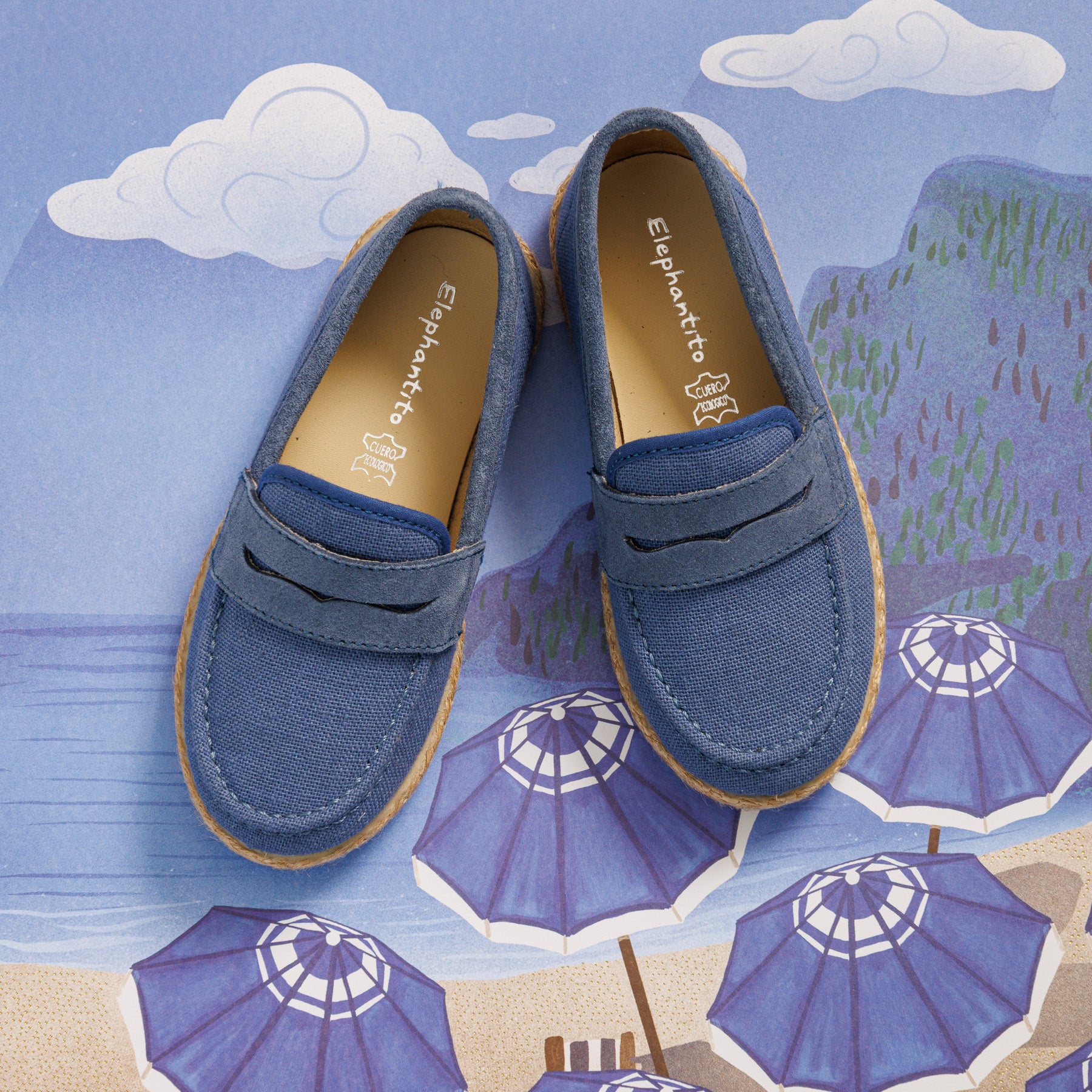 Elephantito - Indigo blue toddler loafer espadrilles with stitched detailing and tan rubber sole on illustrated beach background.
