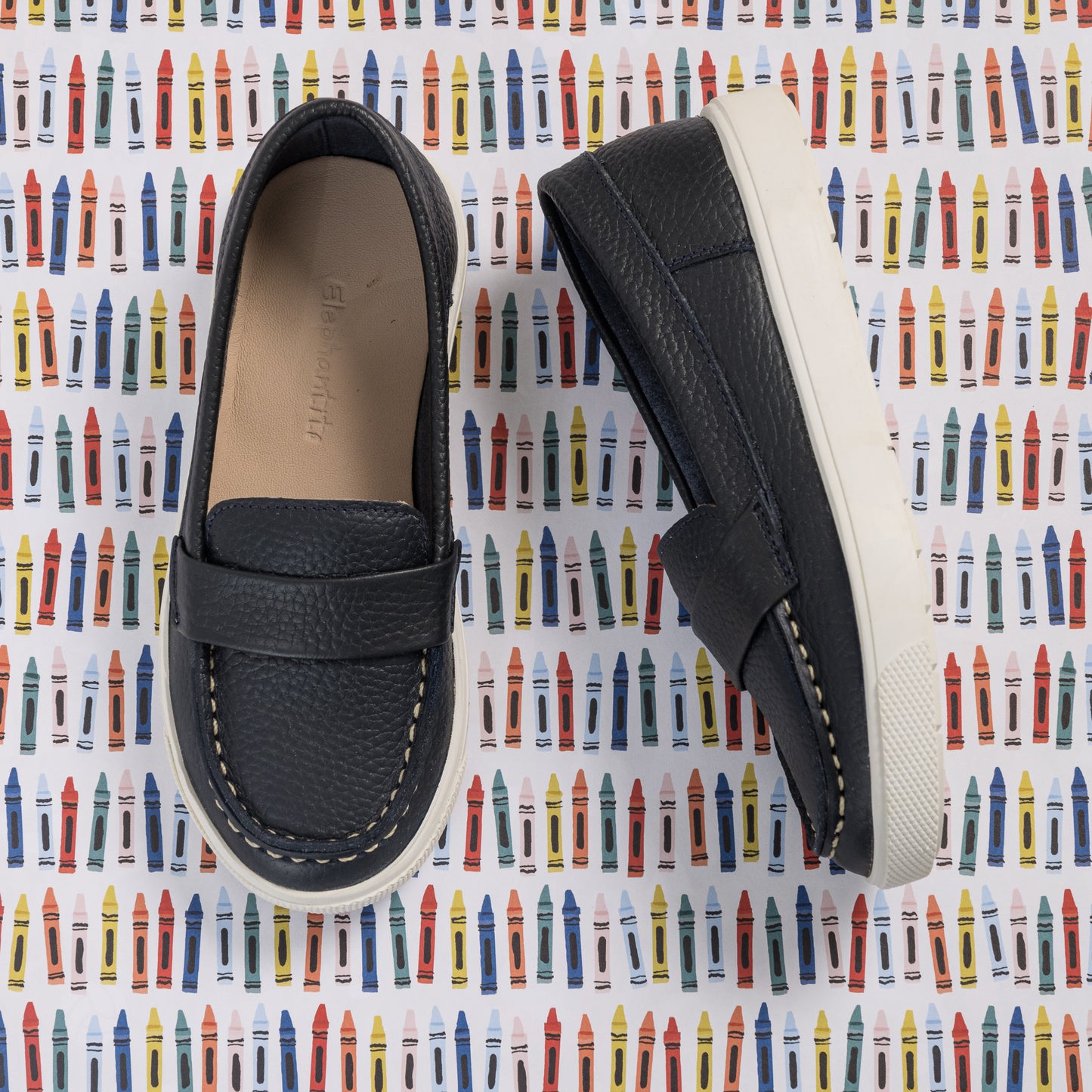 Navy blue leather boys' loafers with white stitching and white rubber soles on patterned background.