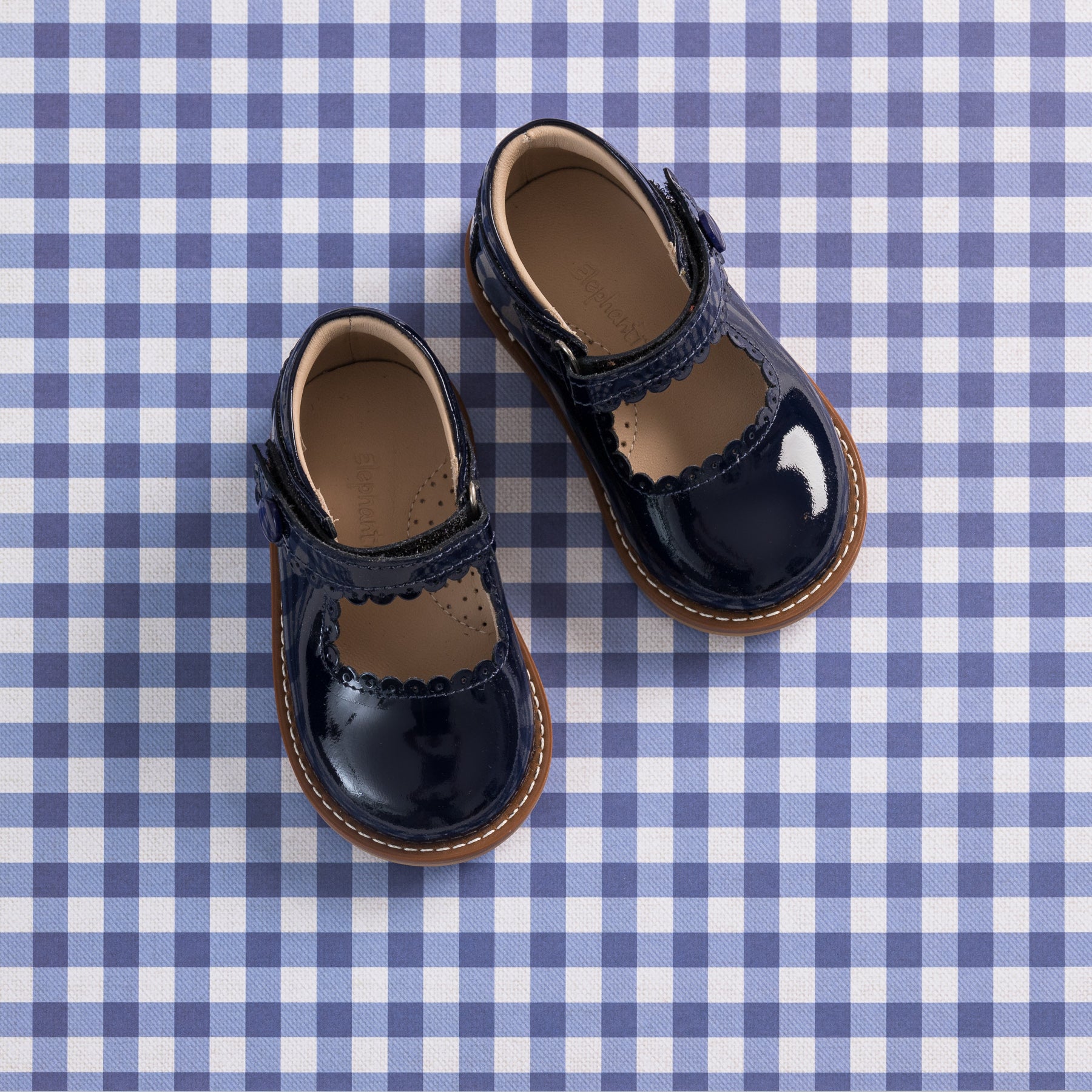 Toddler Mary Jane shoes in glossy navy patent leather with scalloped edges and brown soles on blue gingham background.
