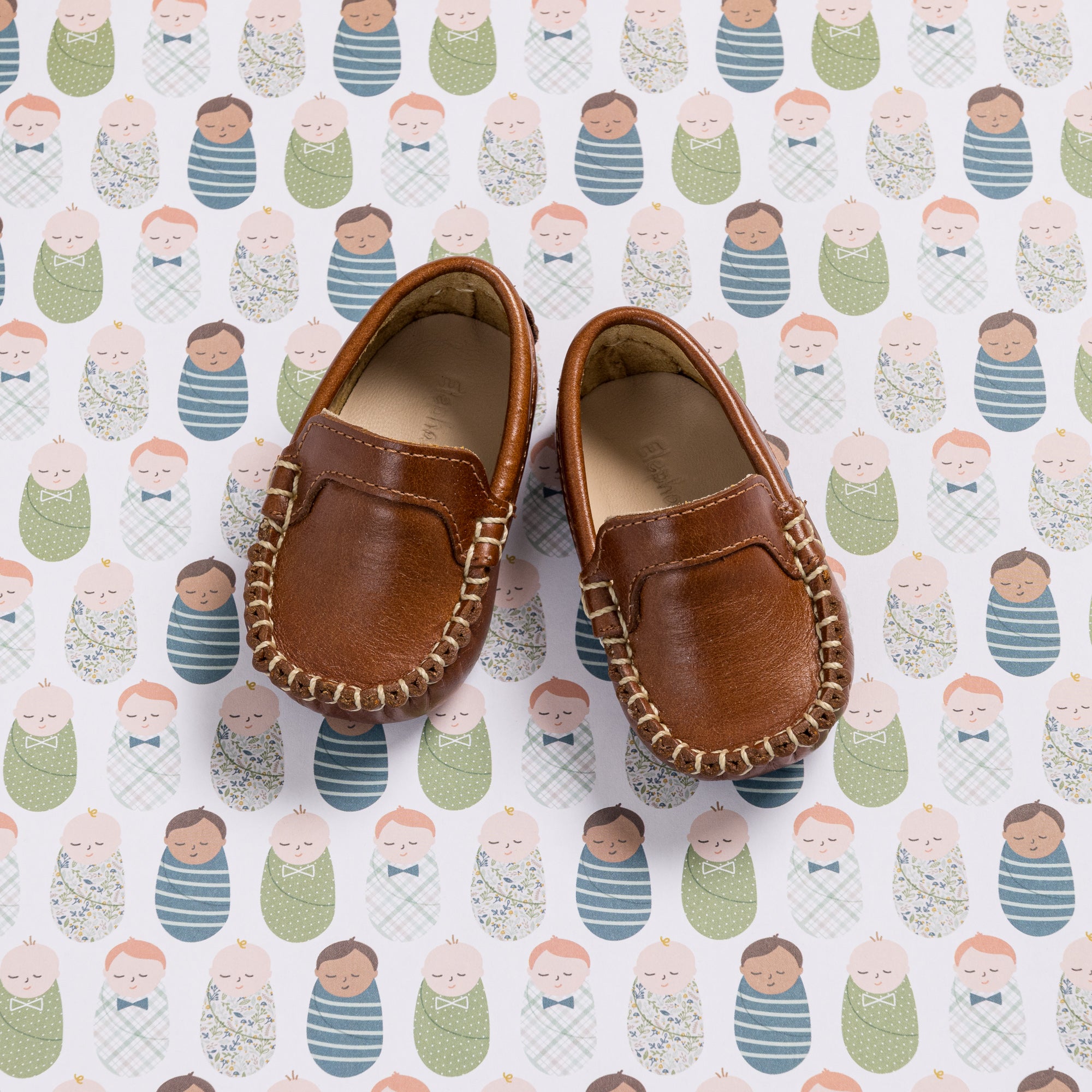 Natural brown leather baby moccasins with stitched detailing and soft soles on patterned background.