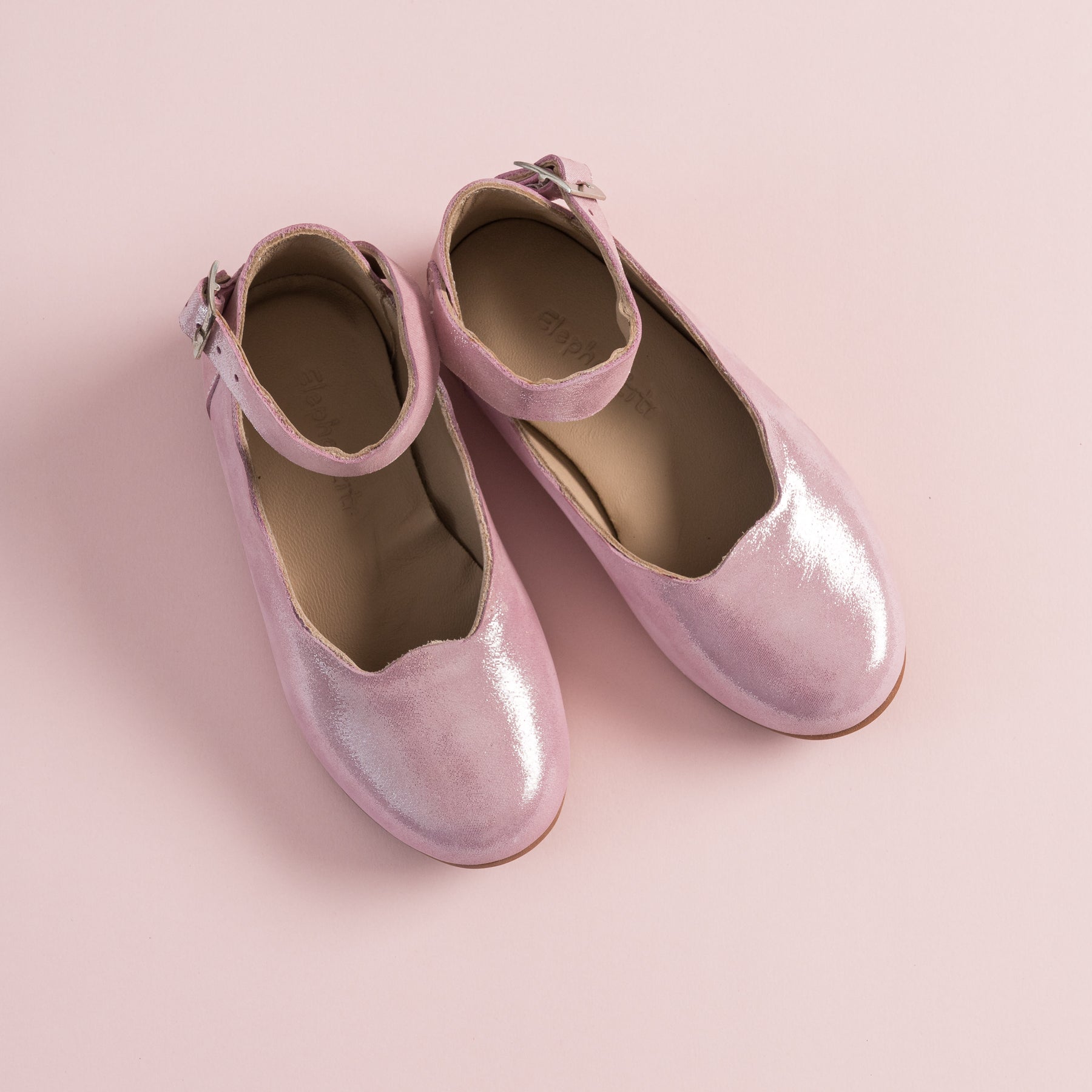 Glossy carnation pink girls' ballet flats with ankle straps and buckle closures on a soft pink background.