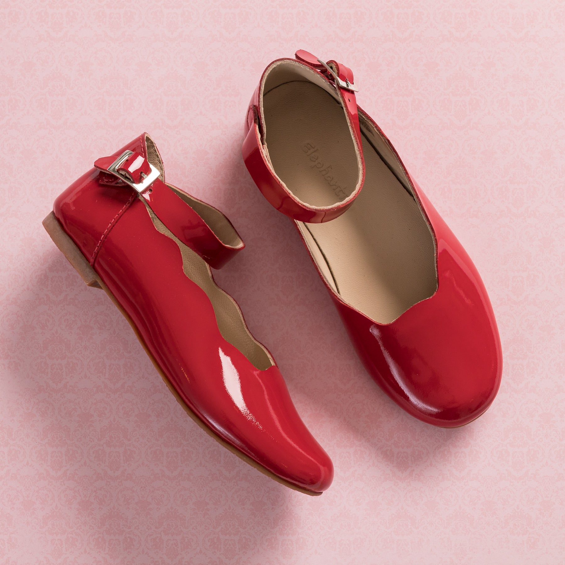 Red patent leather children's flats with scalloped edges and ankle buckle straps on pink background.