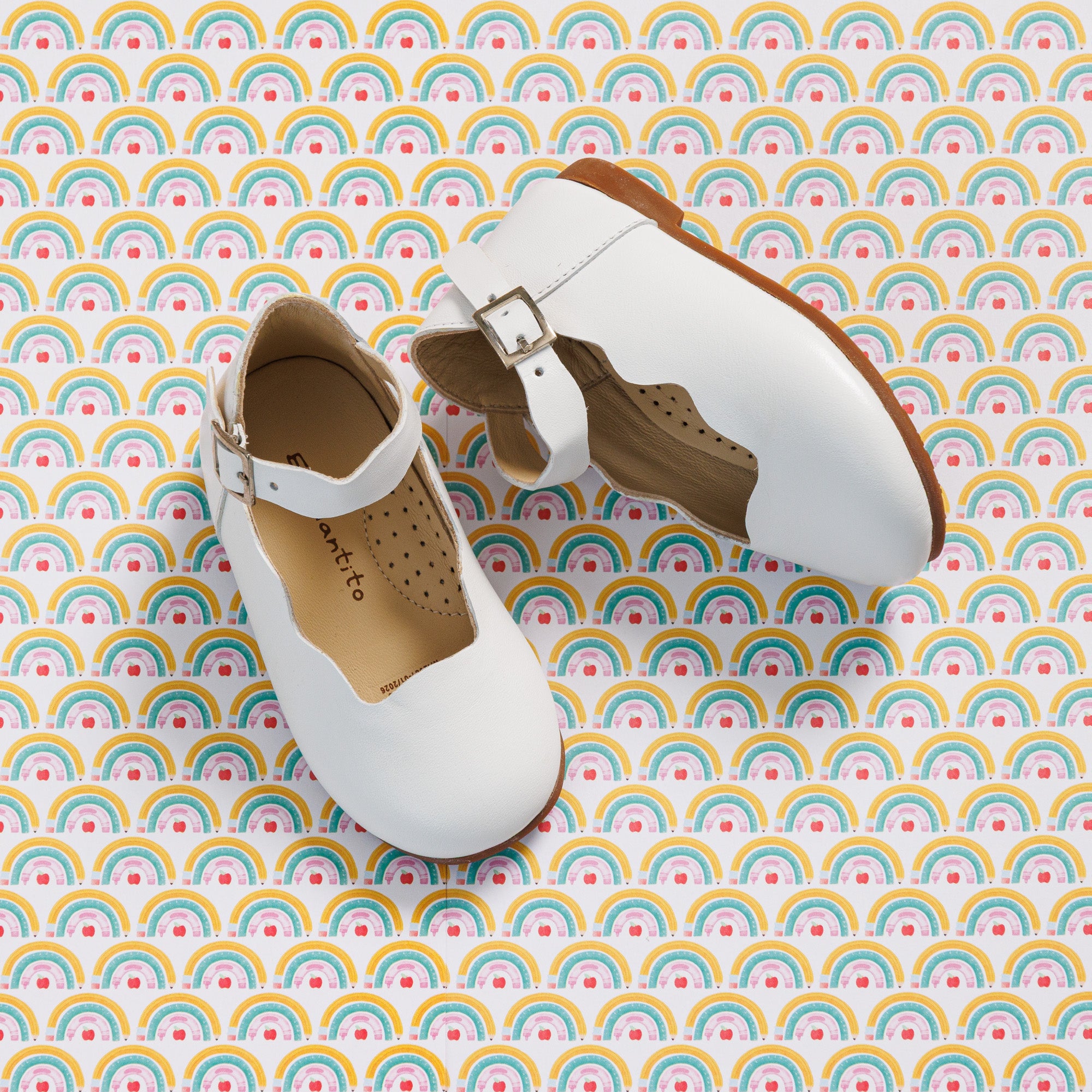Elephantito - White toddler Mary Jane flats with buckle strap and perforated tan interior lining on rainbow-patterned background.
