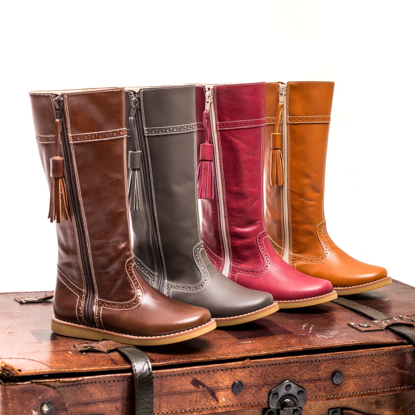 Brown children’s riding boots with tassel detail, rounded toe, and contrast stitching, arranged on vintage suitcase.