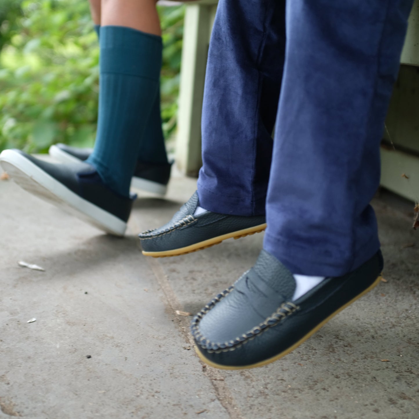 Navy blue children’s loafers with moccasin stitching and rubber soles worn with navy pants and teal socks outdoors.