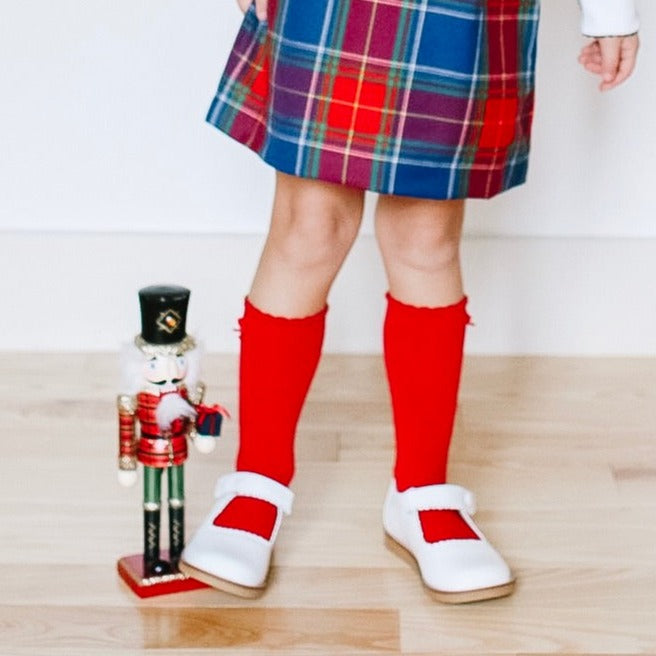 White Mary Jane shoes with red straps worn with red knee-high socks and a plaid skirt.