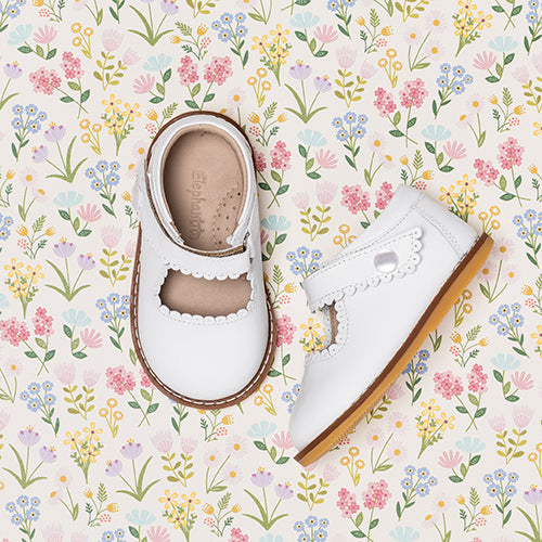 White toddler Mary Jane shoes with scalloped edges and tan rubber soles on floral background.
