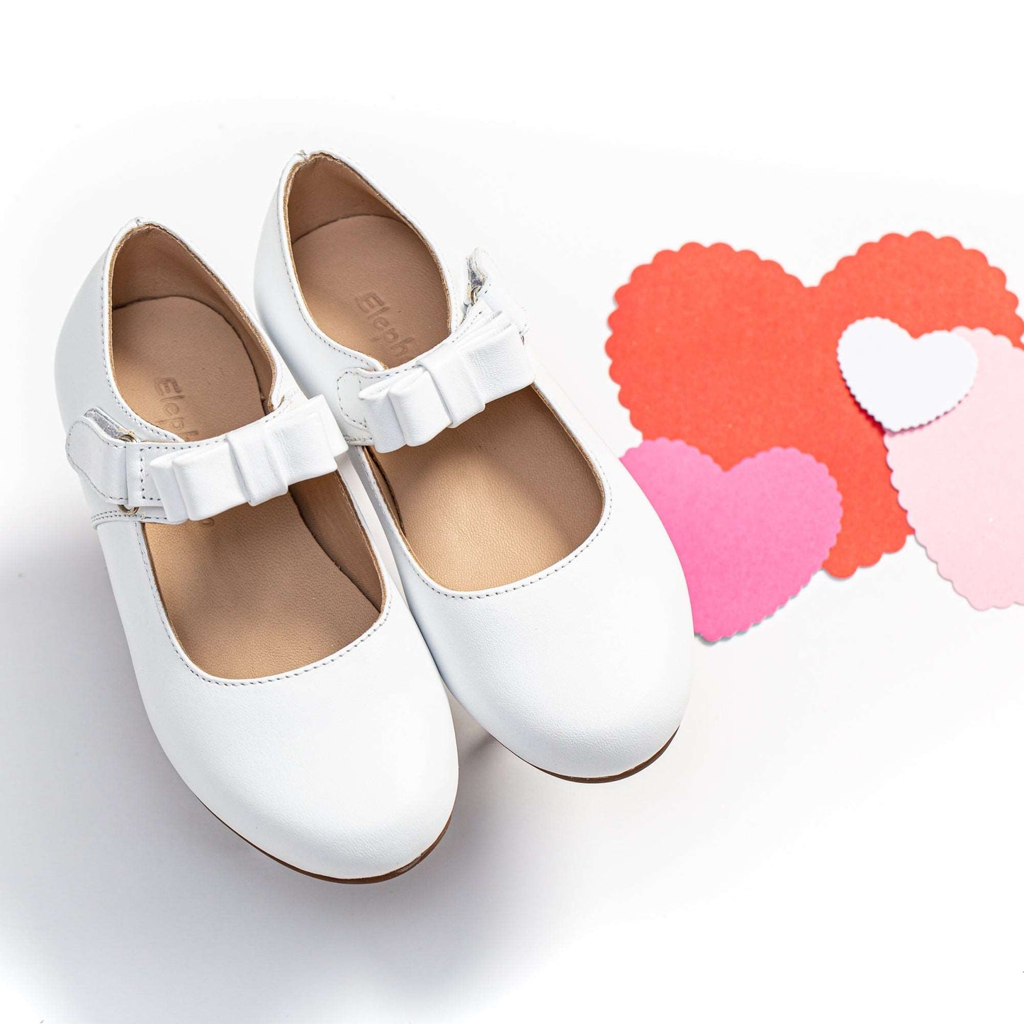 White toddler Mary Jane shoes with bows on adjustable straps next to red and pink heart cutouts.