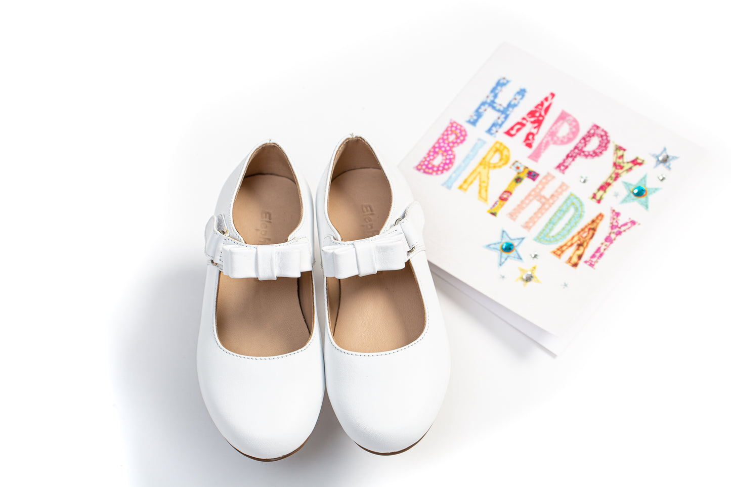 White leather children’s Mary Jane shoes with a single buckle strap, paired with a colorful "Happy Birthday" card.