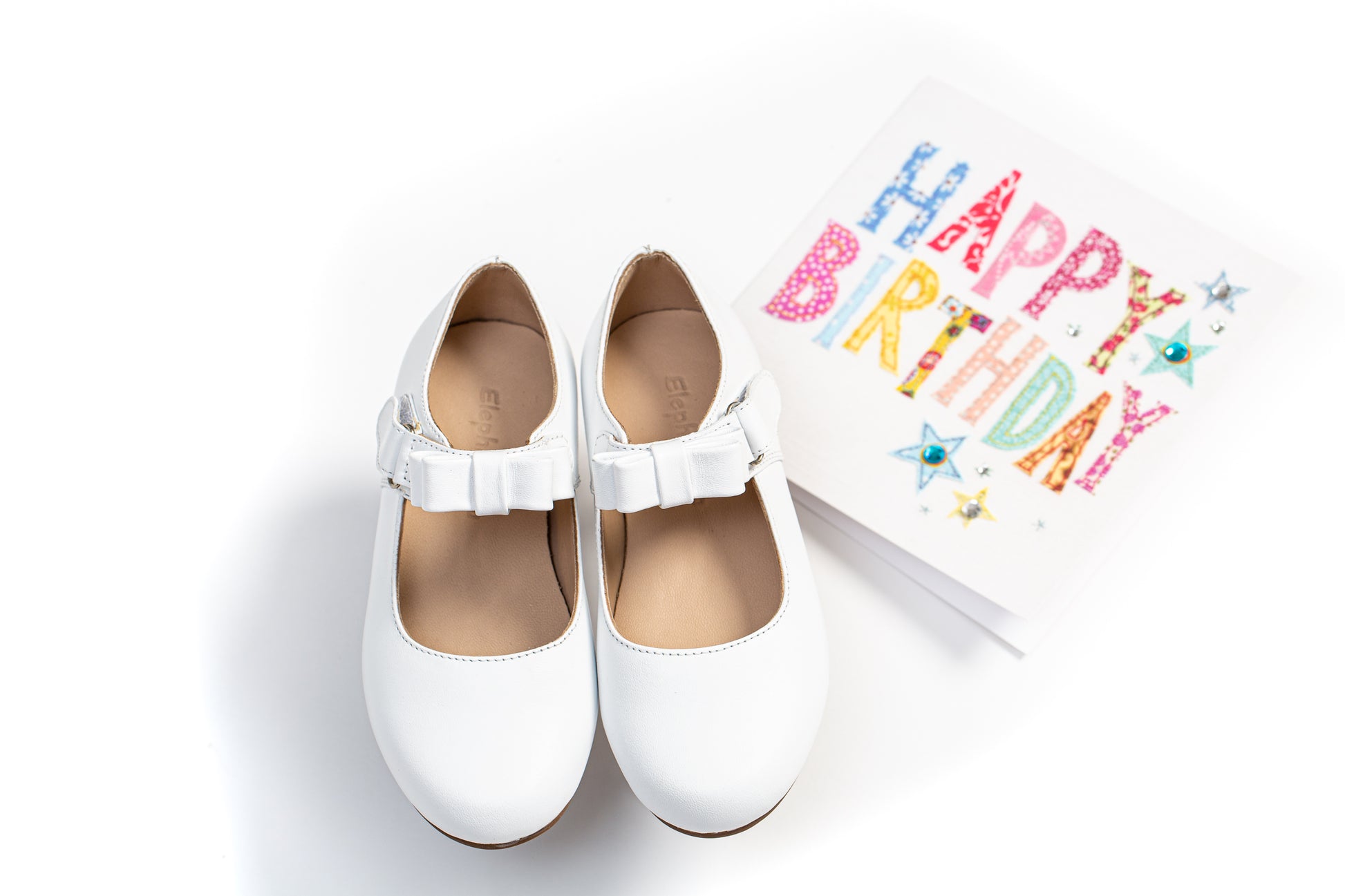White leather children’s Mary Jane shoes with a single buckle strap, paired with a colorful "Happy Birthday" card.