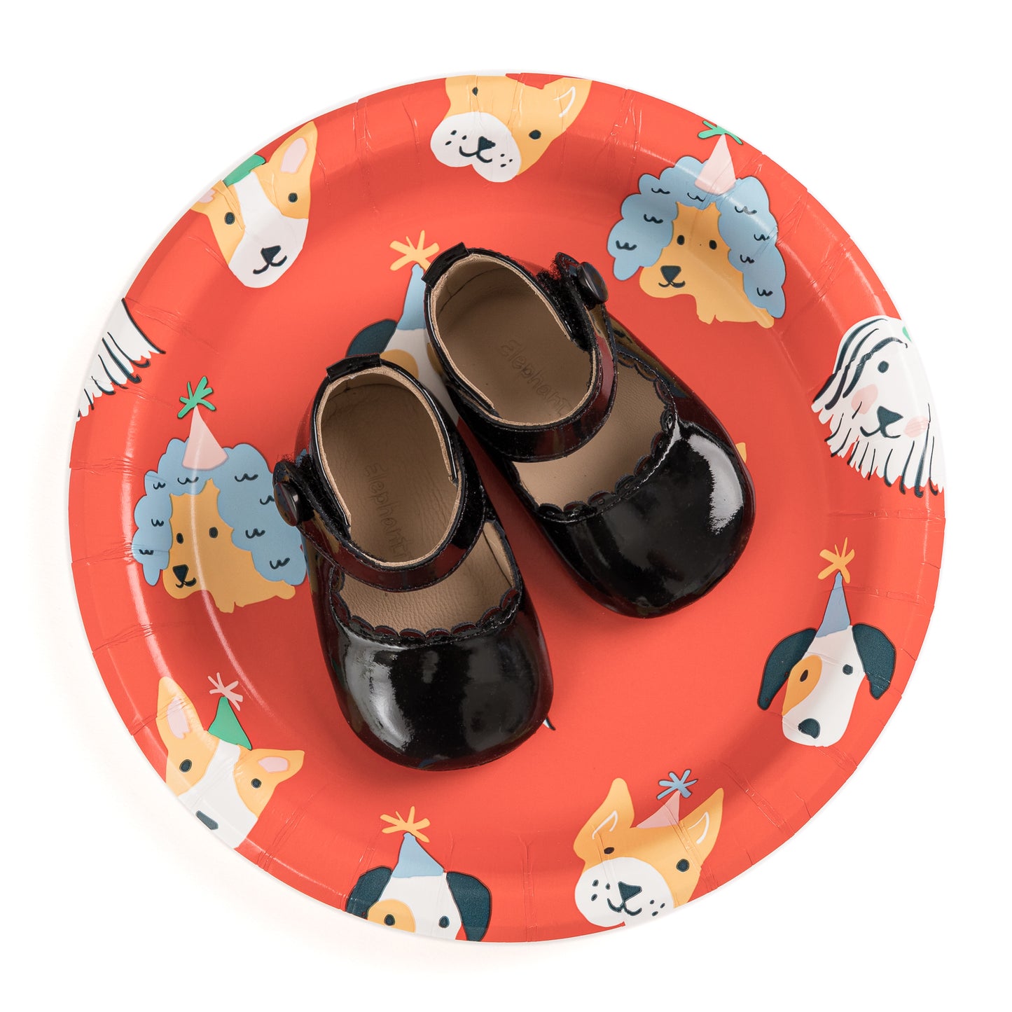 Patent black Mary Jane baby shoes with scalloped edges and ankle straps on a red plate with dog illustrations.