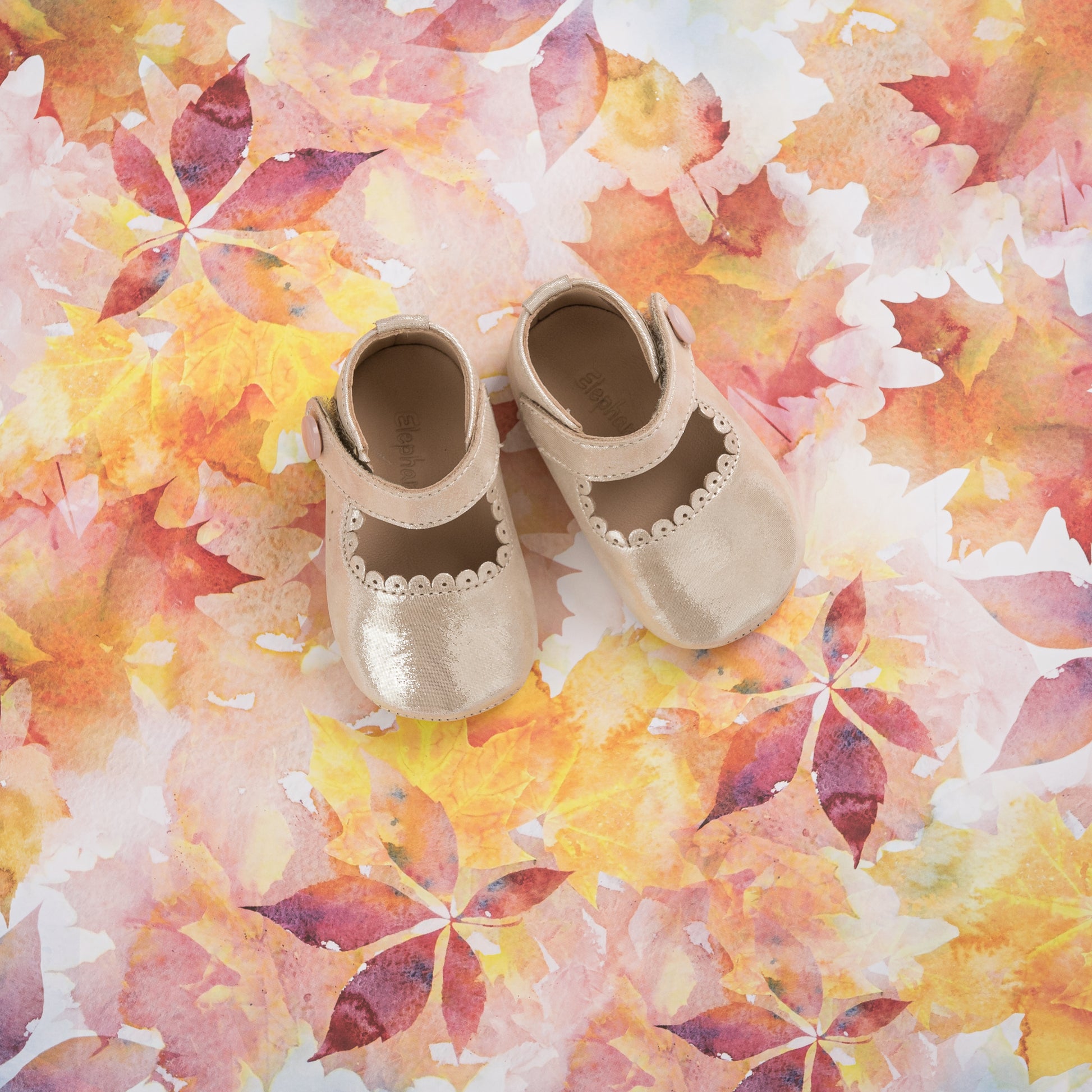 Beige baby Mary Jane shoes with scalloped edges and velcro straps on a colorful autumn leaf background.