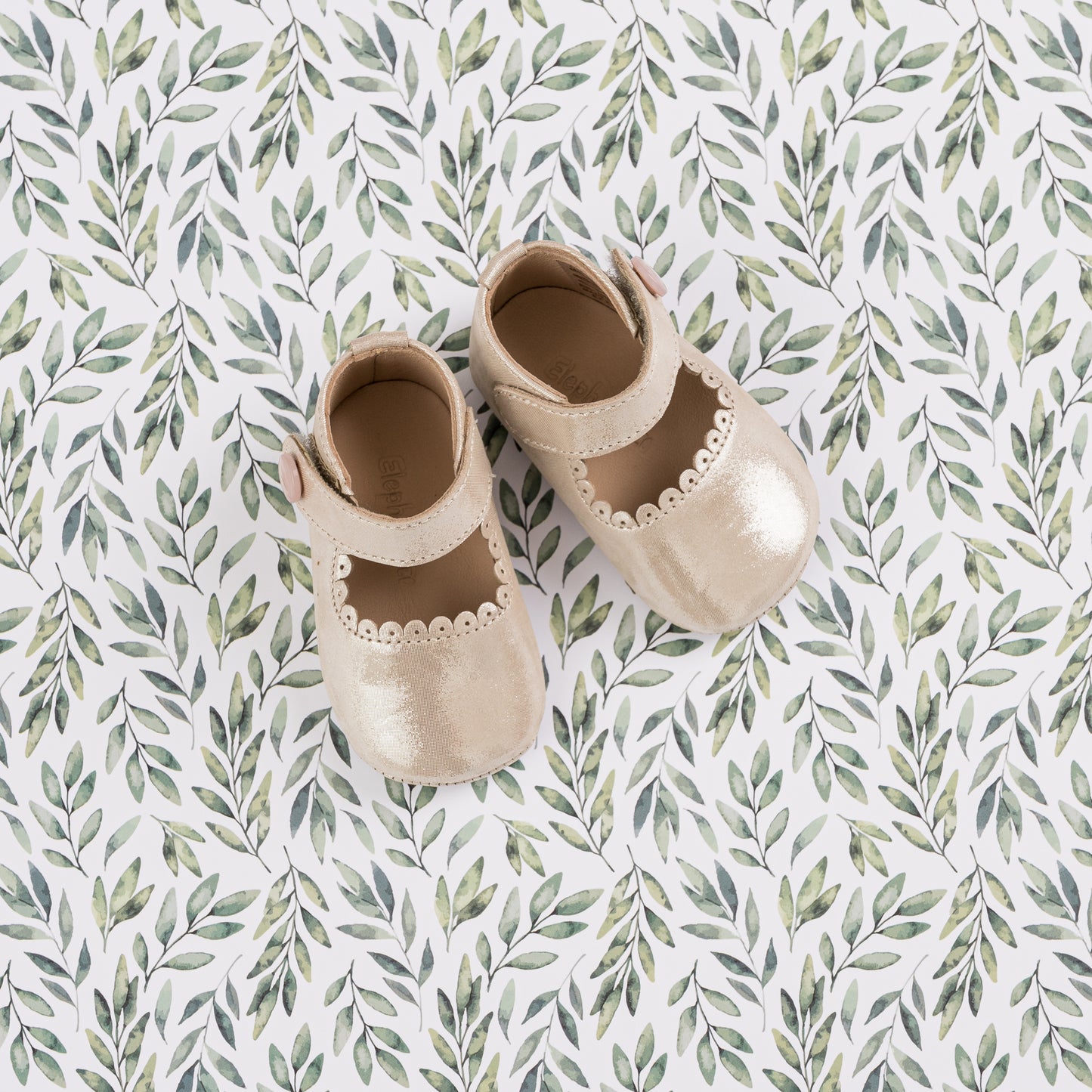 Cream-colored baby Mary Jane shoes with scalloped edges and soft leather material on a leafy patterned background.