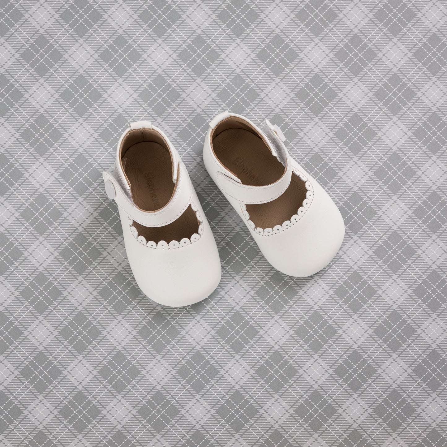 White baby Mary Jane shoes with scalloped edge and Velcro strap on gray plaid background.