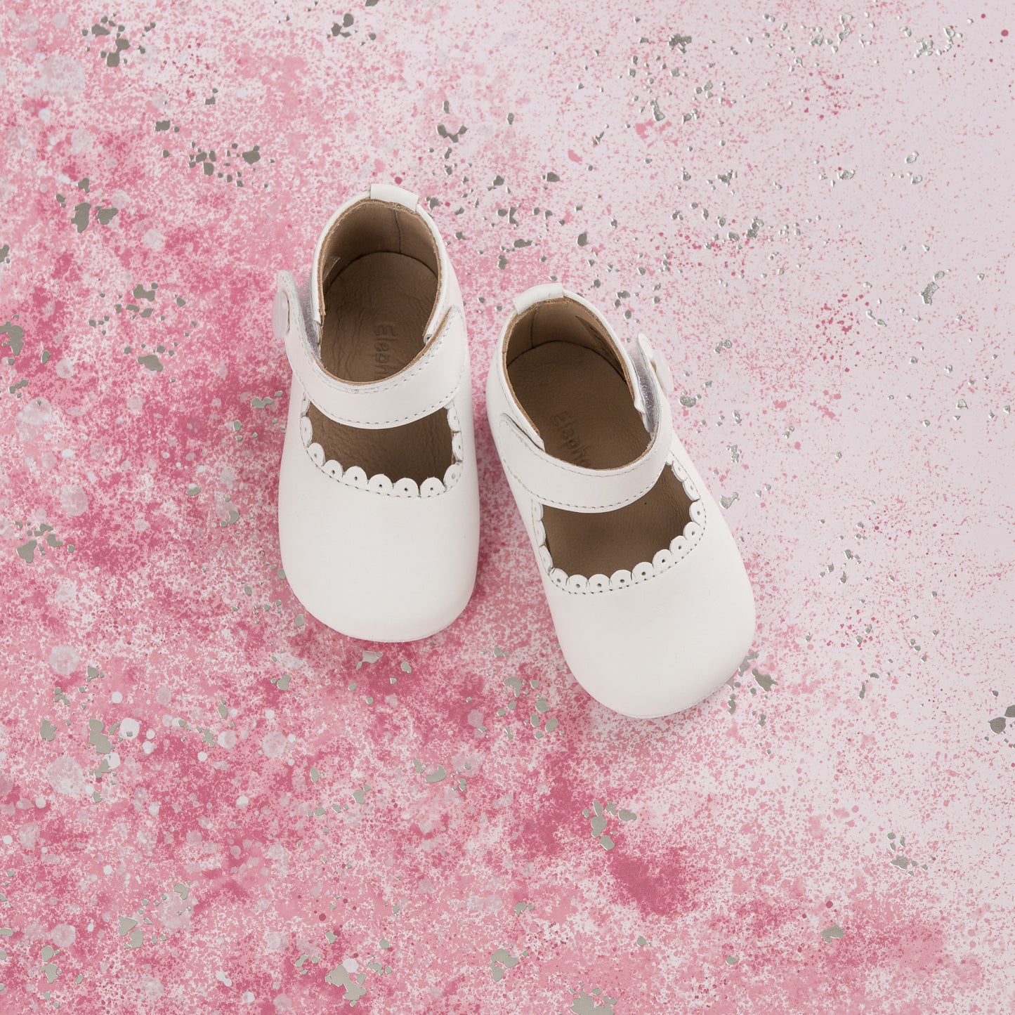 White baby Mary Jane shoes with scalloped edges and soft leather material on pink textured background.