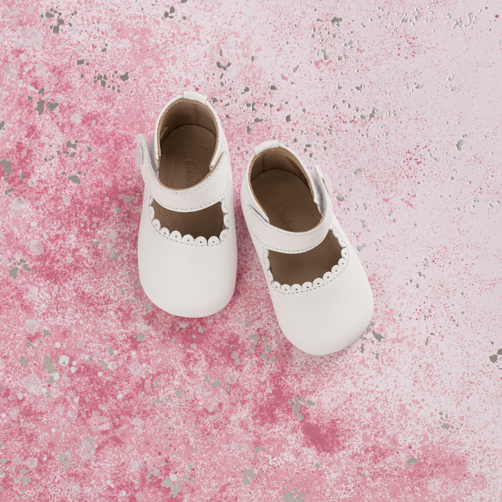 White baby Mary Jane shoes with scalloped edges and soft leather material on pink textured background.