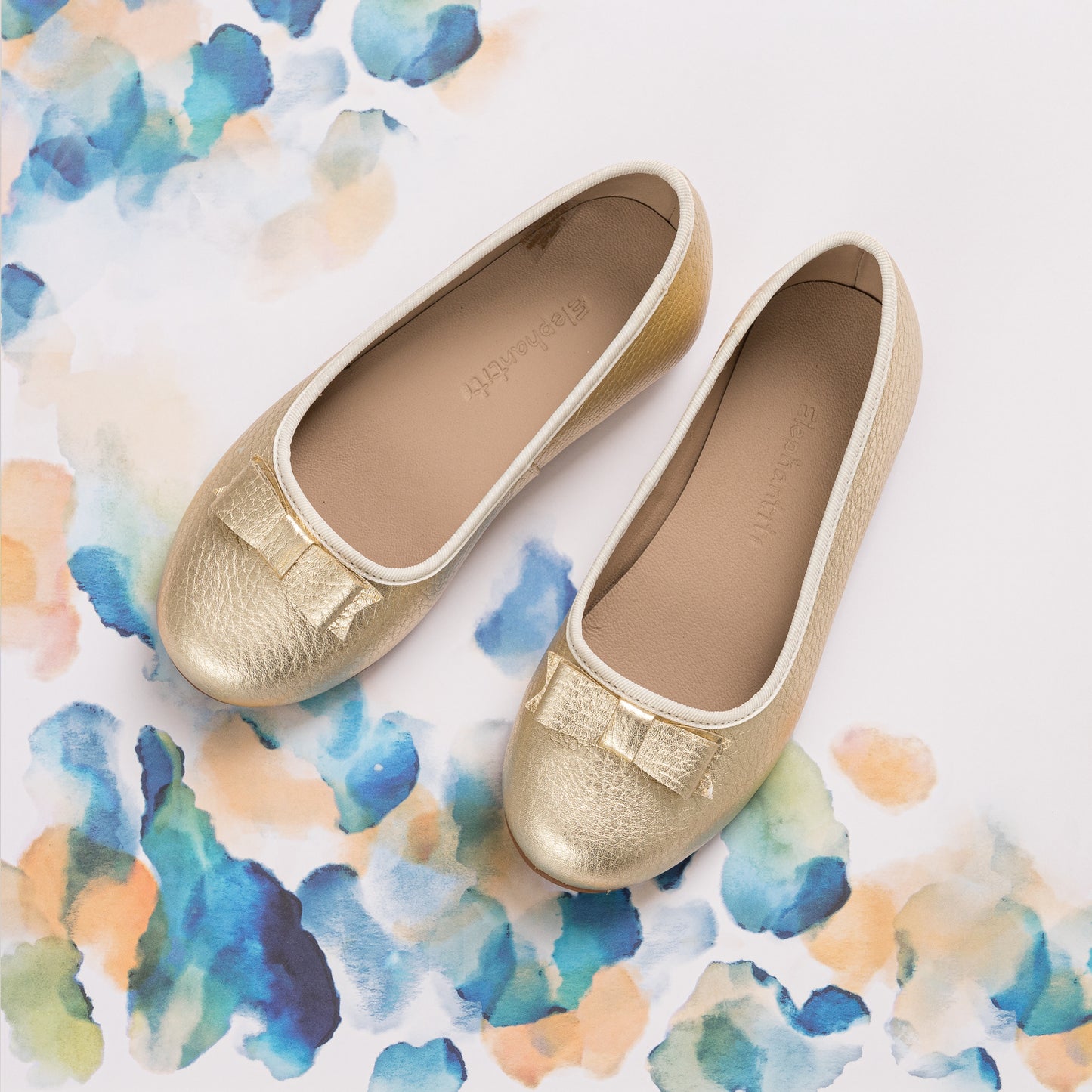 Gold metallic toddler ballet flats with small bows and white trim on a watercolor background.