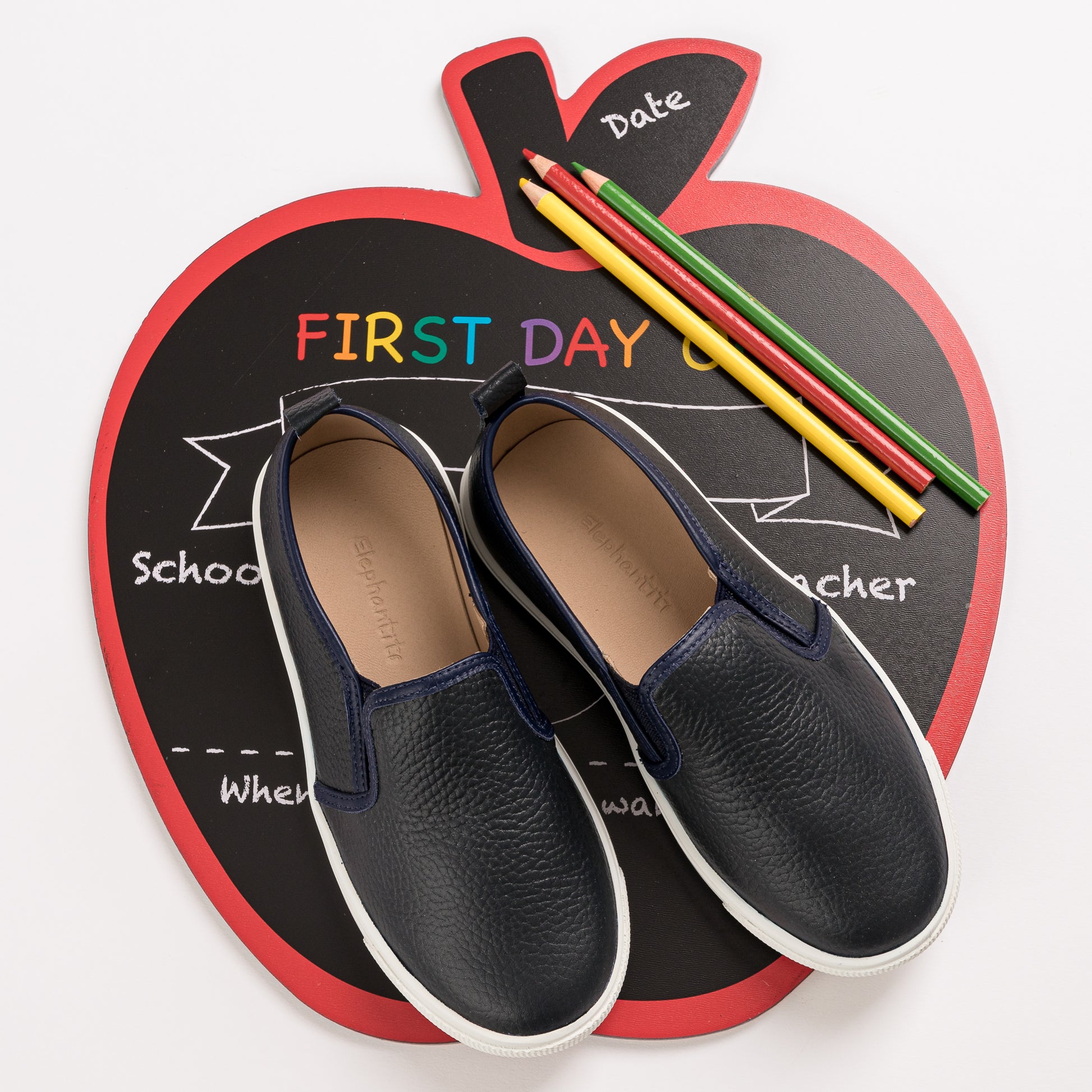 Textured navy blue slip-on shoes with white soles for children on apple-shaped chalkboard background with colored pencils.