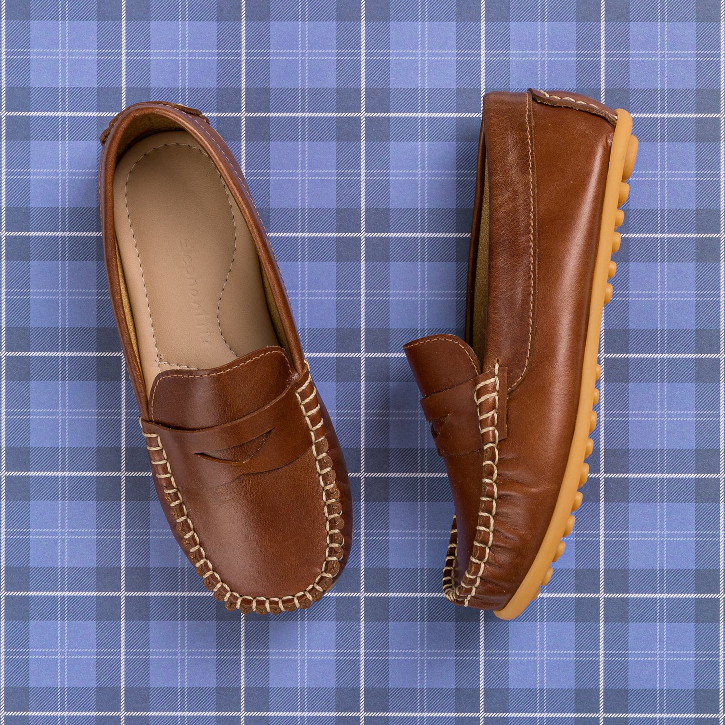 Natural brown leather boys loafer with moccasin stitching and tan rubber sole on blue plaid background.