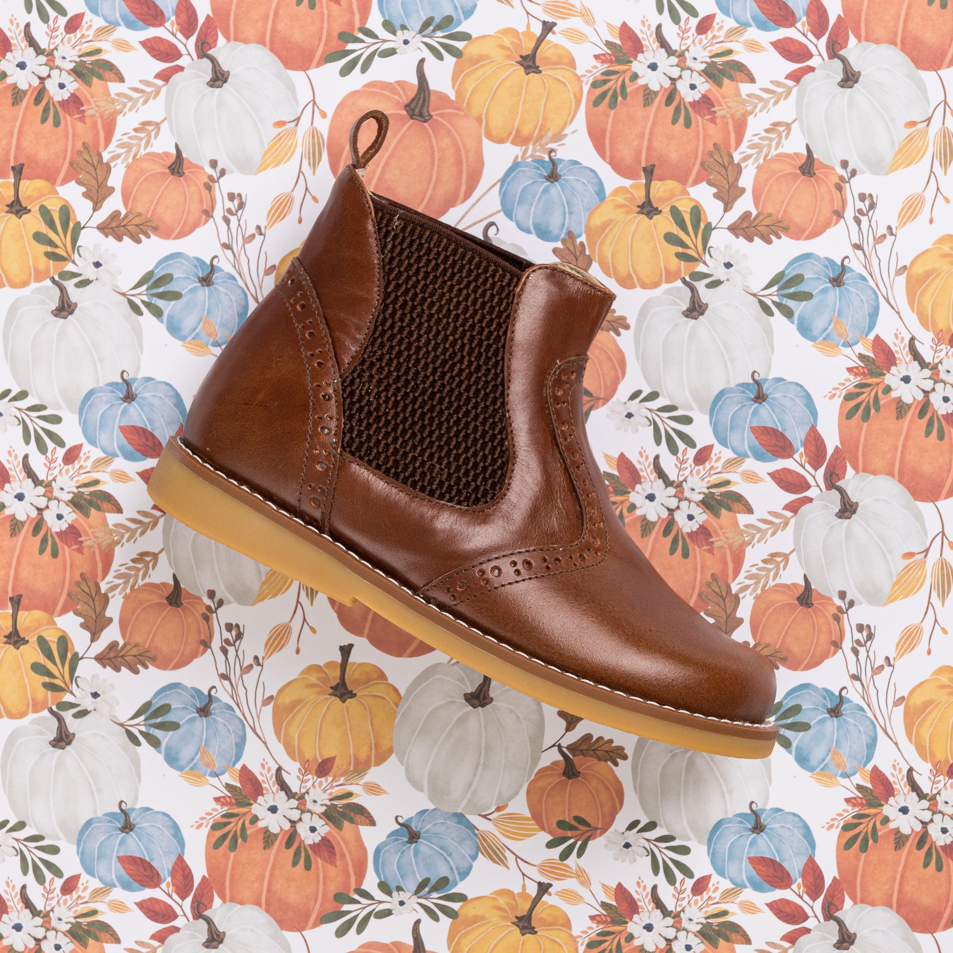 Brown leather children's bootie with elastic side panels and pull tab on a floral pumpkin background.