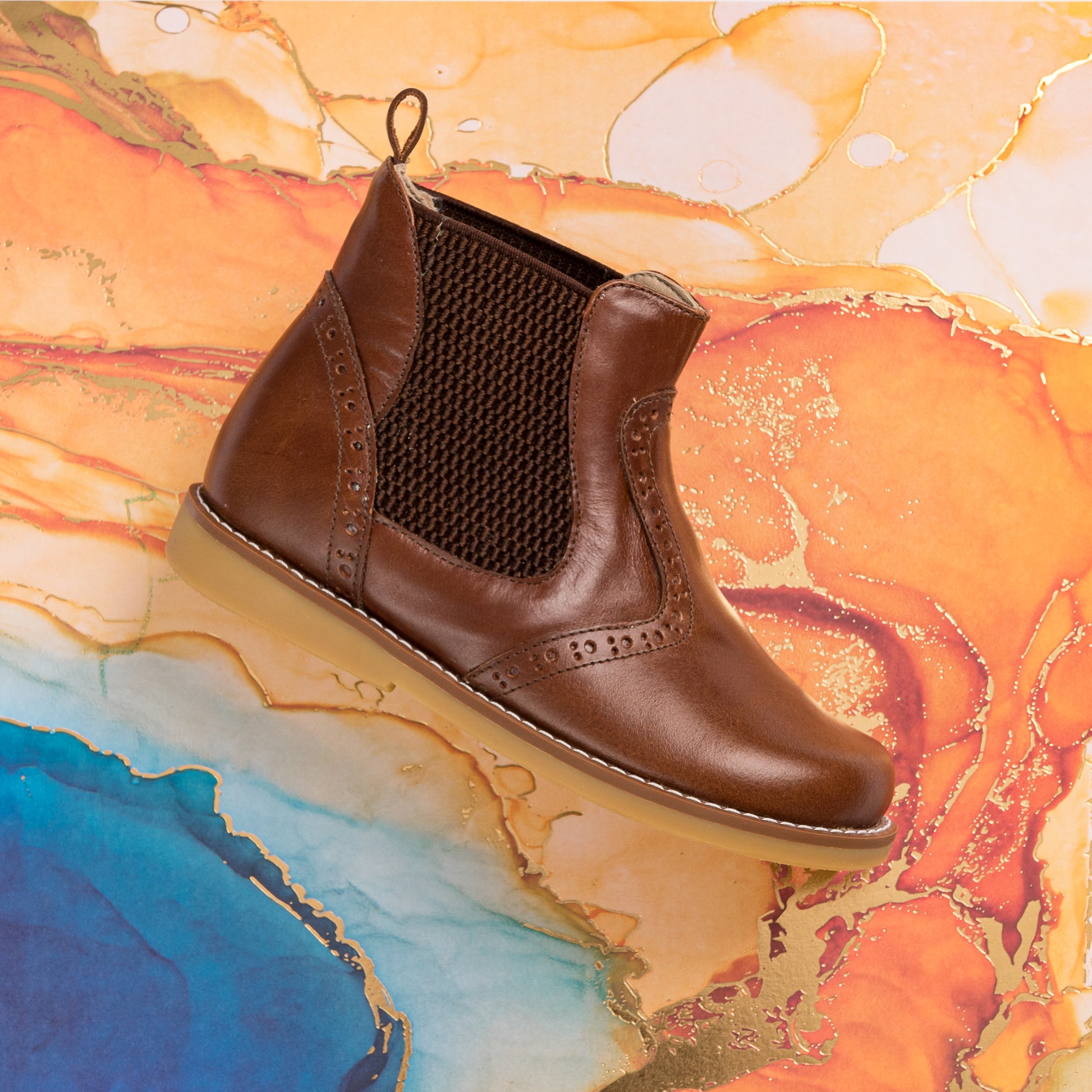 Brown leather children's bootie with elastic side panels and tan rubber sole on colorful marbled background.
