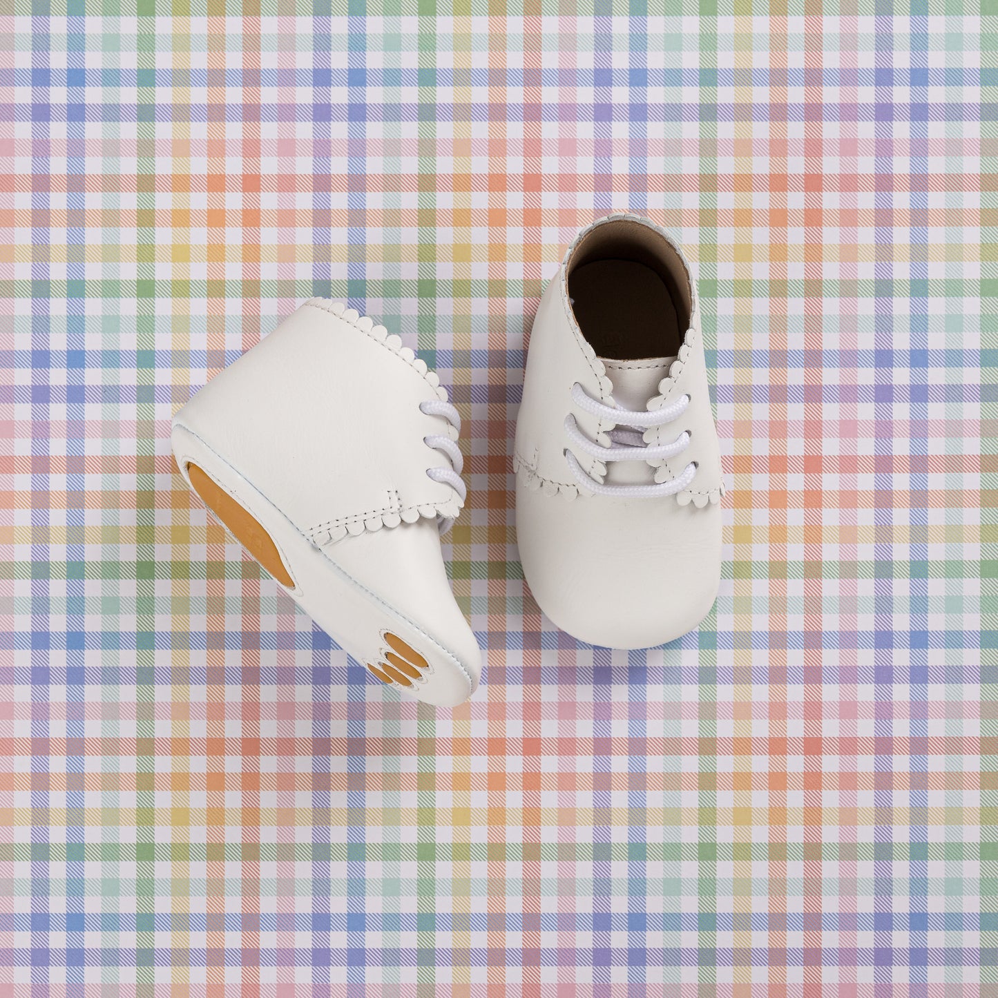 White infant booties with scalloped edges and lace-up design on pastel plaid background.