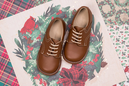 Brown leather toddler shoes with white stitching and tan laces on festive floral fabric background.