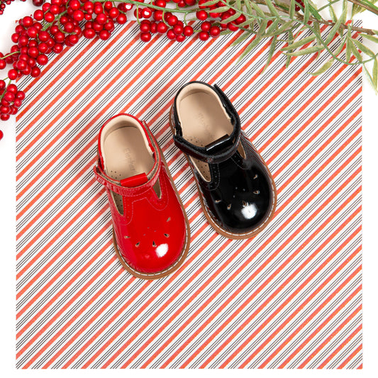 Patent leather T-bar toddler shoes in red and black on striped background with holiday berries and greenery.
