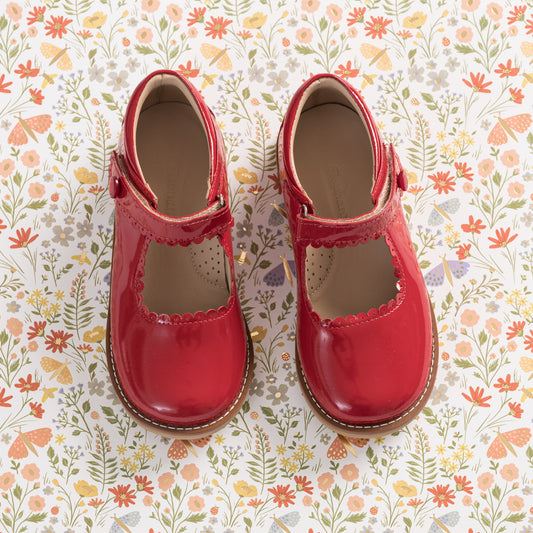 Glossy red patent leather Mary Jane shoes with scalloped edges and a single strap buckle on floral background.