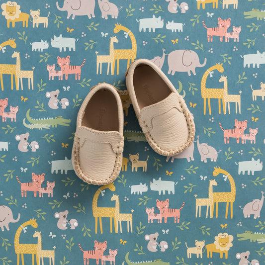 Cream-colored baby moccasins with soft leather and stitched detailing on a blue animal print mat.