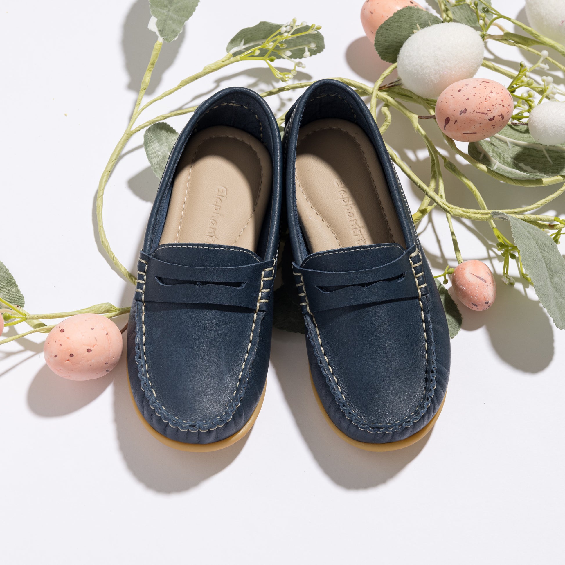 Navy blue toddler moccasins with stitched detailing and cushioned insoles placed on light background with decorative eggs.