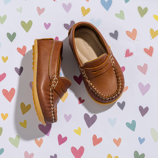 Tan leather toddler moccasins with fringe detail and stitched saddle design on heart-patterned background.