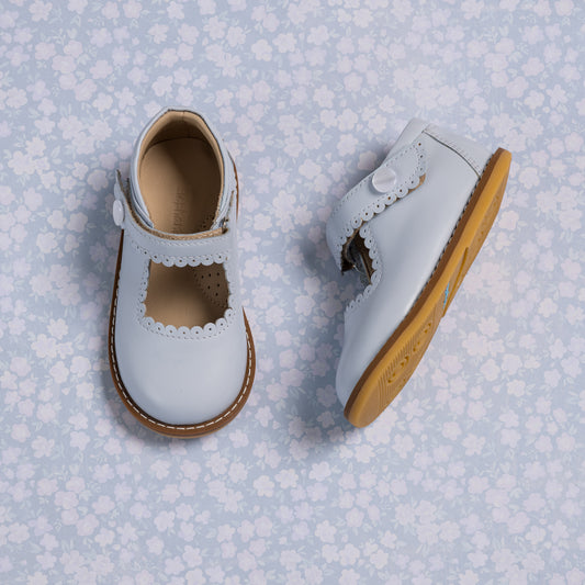 Light blue toddler Mary Jane shoes with scalloped edges and tan soles on floral background.