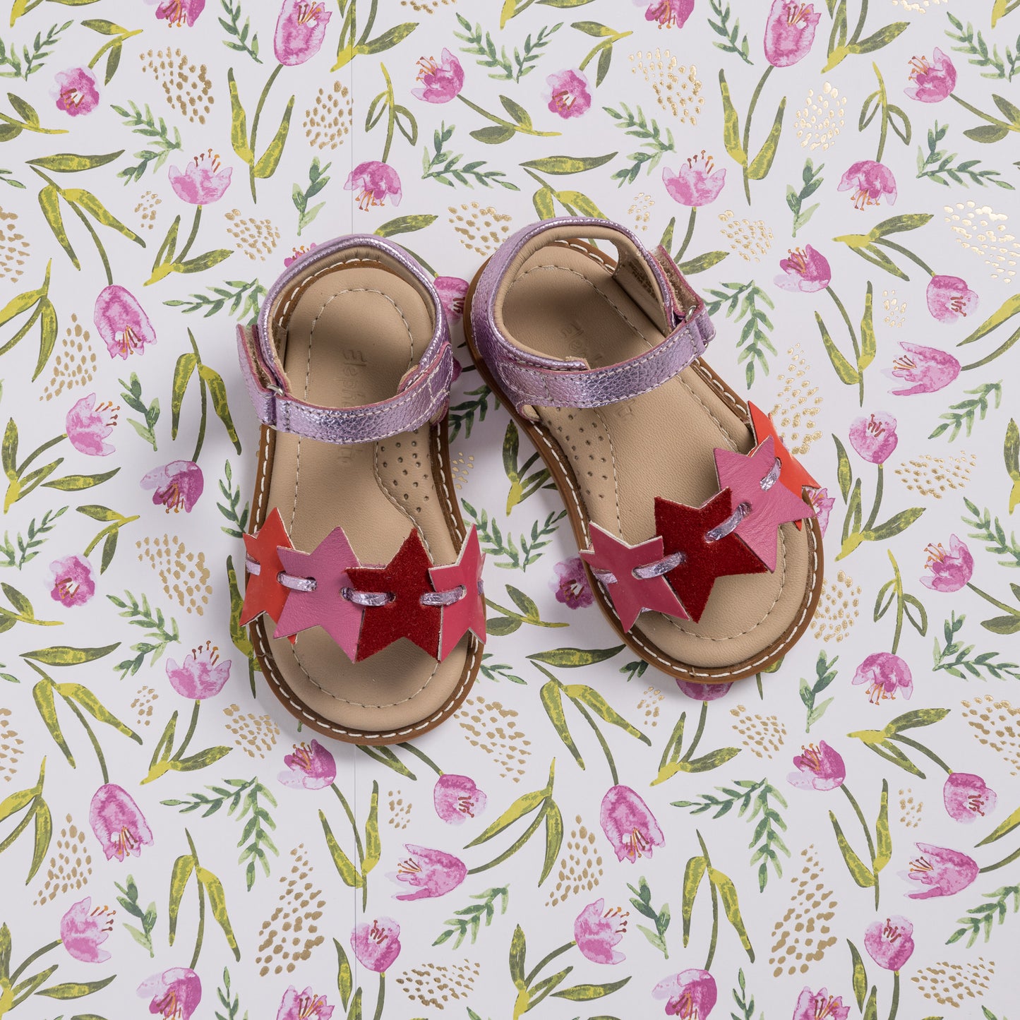 Toddler sandals with pink and fuchsia star-shaped straps and beige soles on floral background.