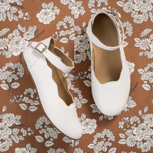 White leather children’s ballet flats with ankle strap and scalloped edge on floral fabric background.