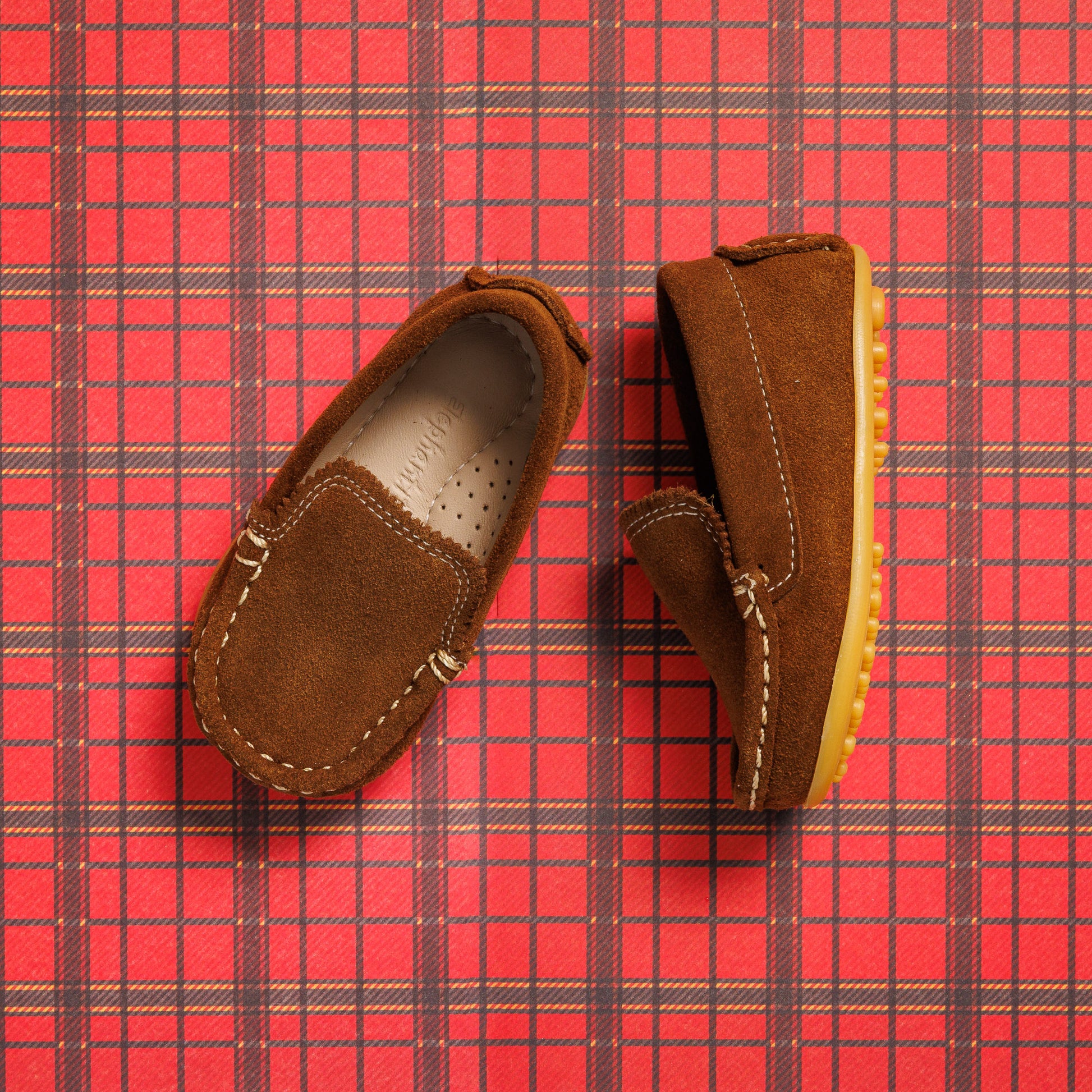 Chestnut suede toddler moccasin driver shoes with contrast yellow stitching and rubber sole on red plaid background.