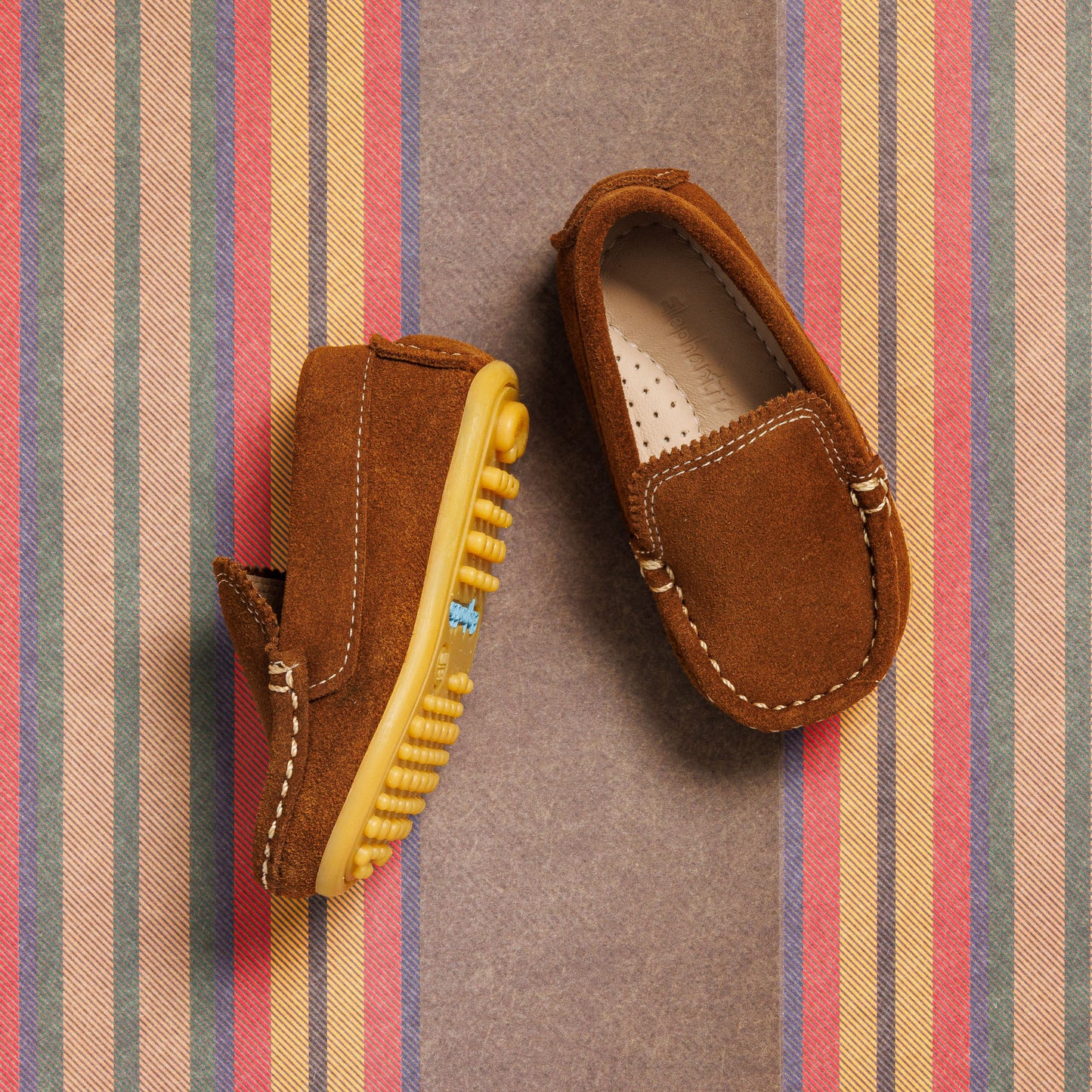 Chestnut suede toddler moccasins with yellow rubber sole and contrast stitching on striped background.