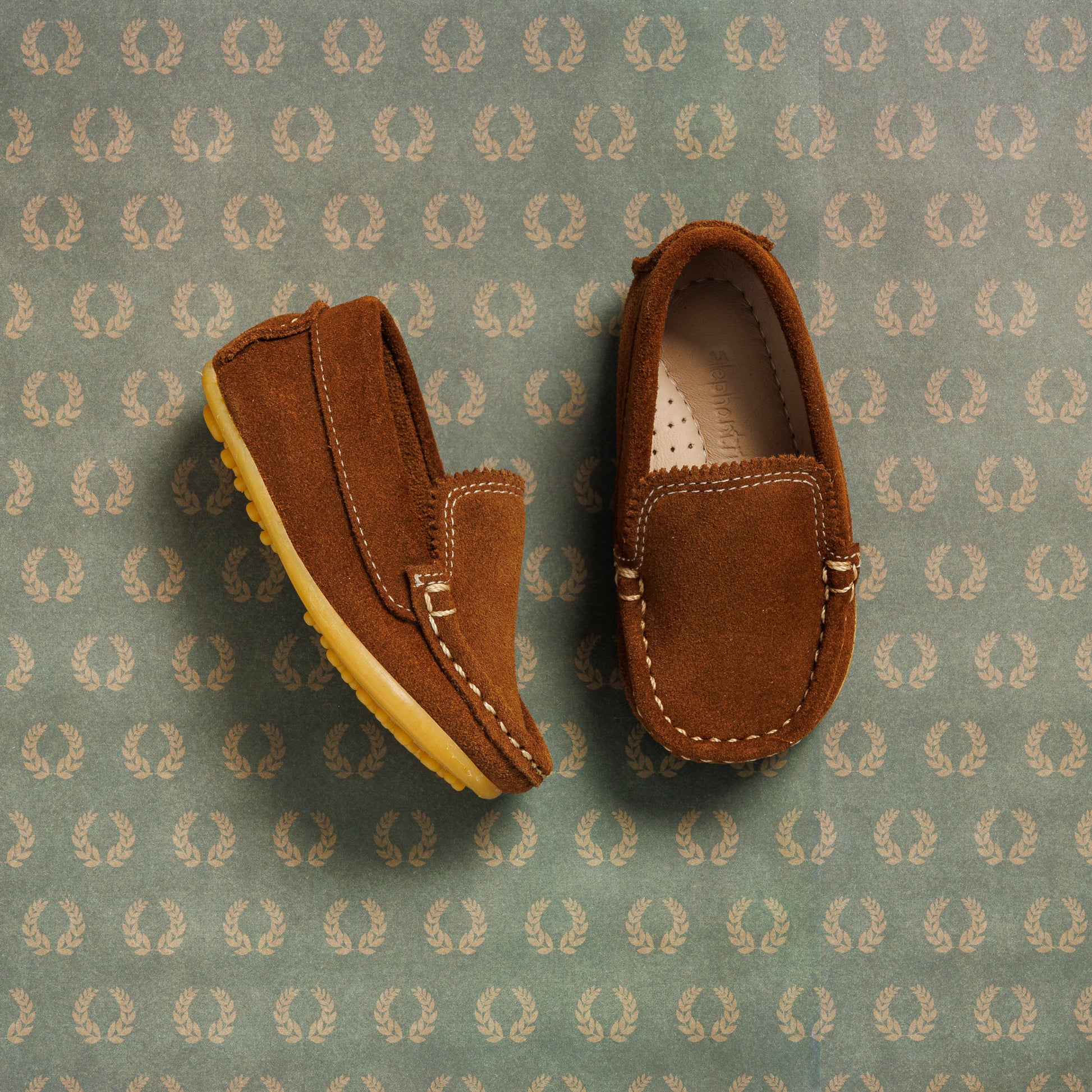 Chestnut suede moccasin toddler shoes with stitched details and rubber soles on patterned background.