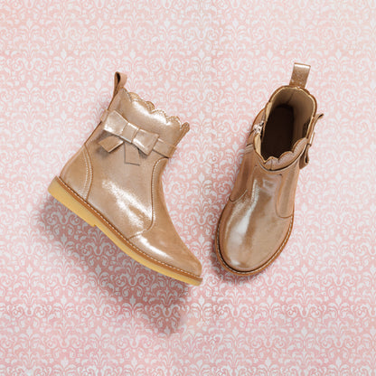 Shiny blush pink toddler booties with scalloped trim and side bow detail on tan soles.