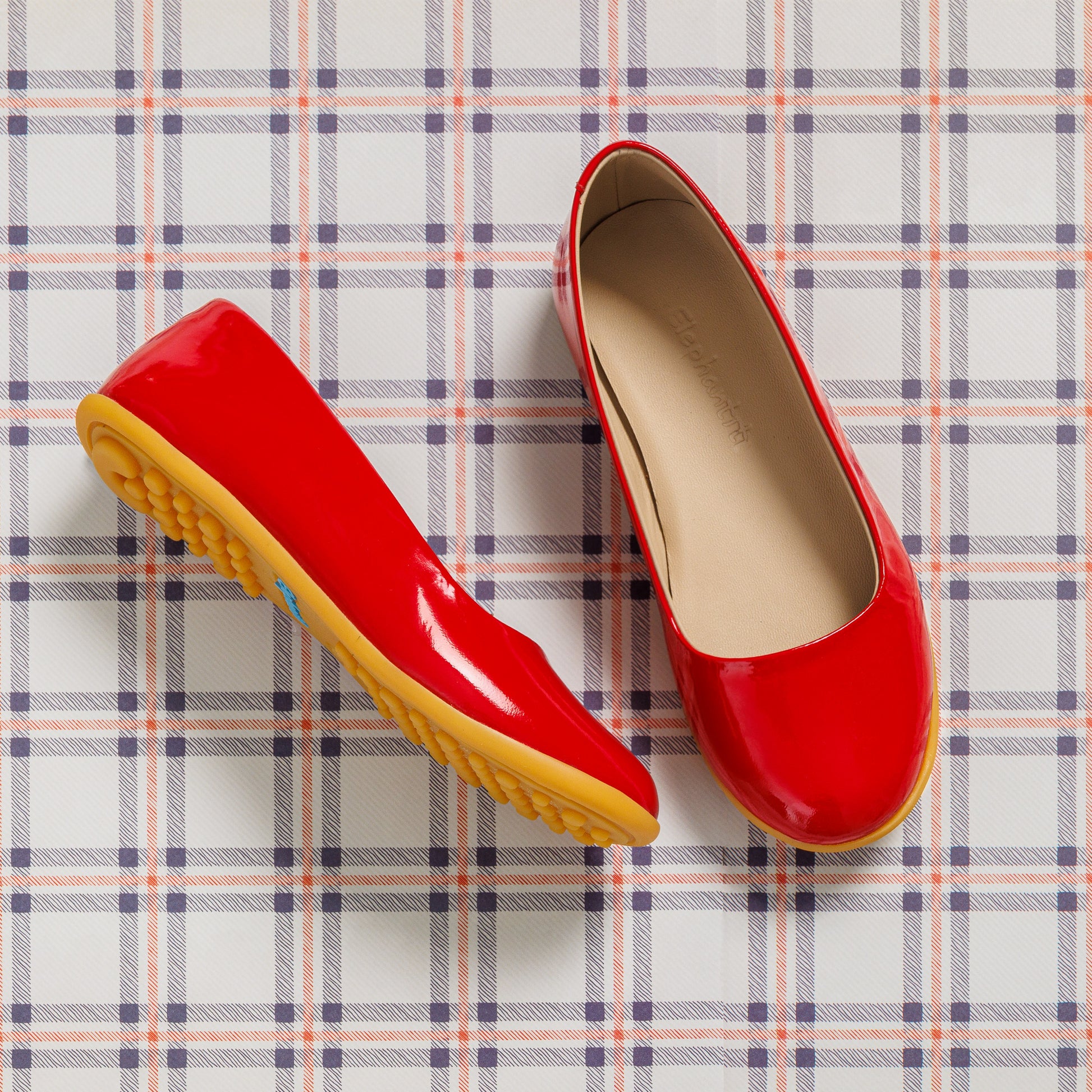 Shiny red toddler ballerina flats with tan cushioned insoles and flexible yellow rubber soles on plaid background.