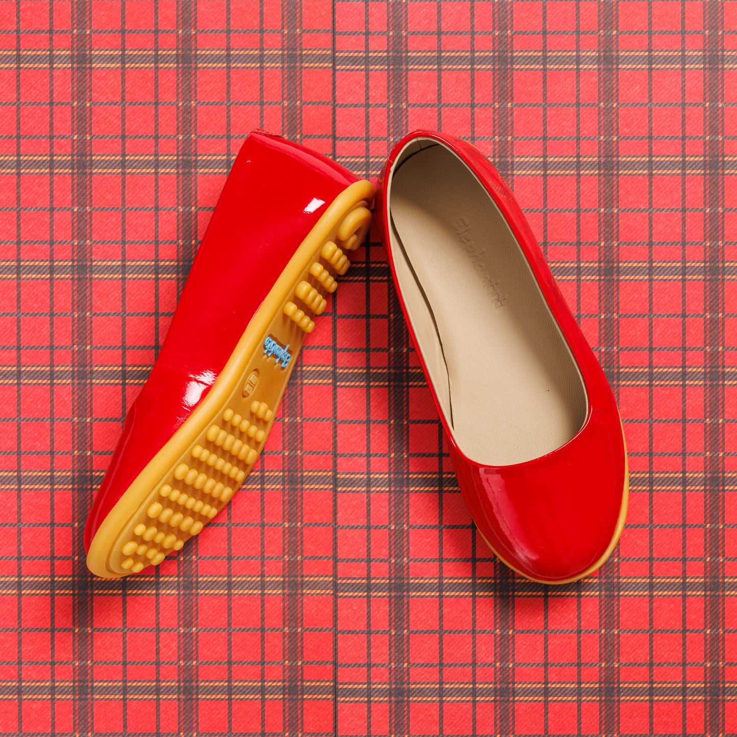 Glossy red toddler ballerina flats with soft beige insoles and textured tan rubber soles on red plaid background.