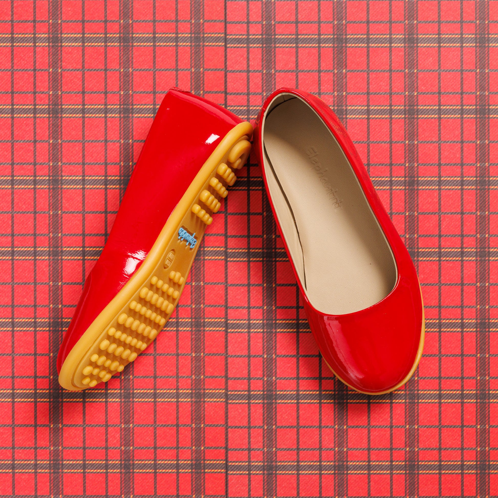 Glossy red toddler ballerina flats with soft beige insoles and textured tan rubber soles on red plaid background.