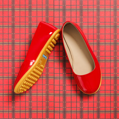 Glossy red toddler ballerina flats with soft beige insoles and textured tan rubber soles on red plaid background.