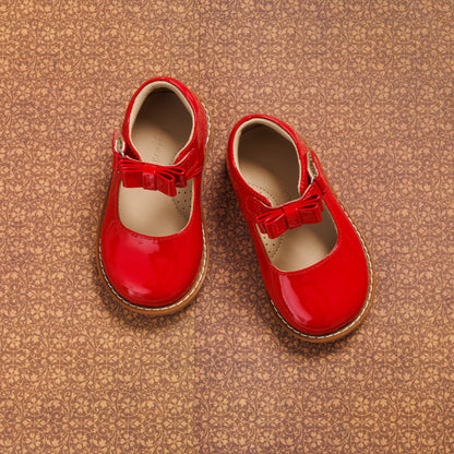 Glossy red toddler Mary Jane shoes with a bow detail and brown soles on a textured background.