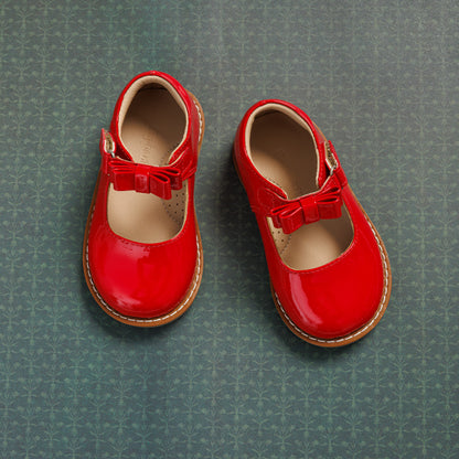 Shiny red toddler Mary Jane shoes with a bow detail and tan sole on green patterned background.
