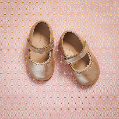 Blush pink toddler Mary Jane shoes with scalloped edges and velcro straps on patterned pink background.