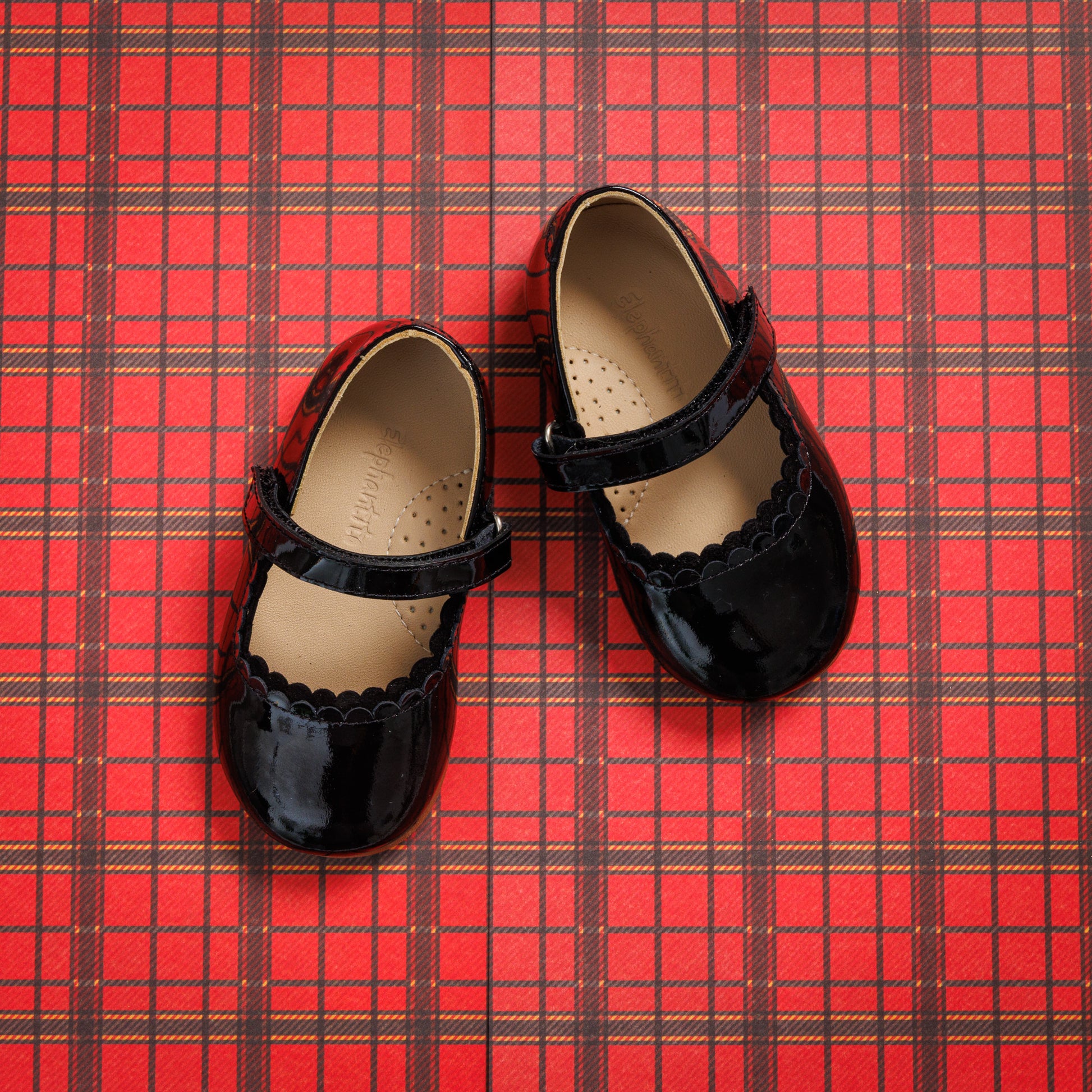 Black patent leather toddler Mary Jane flats with scalloped edges and velcro straps on red plaid background.