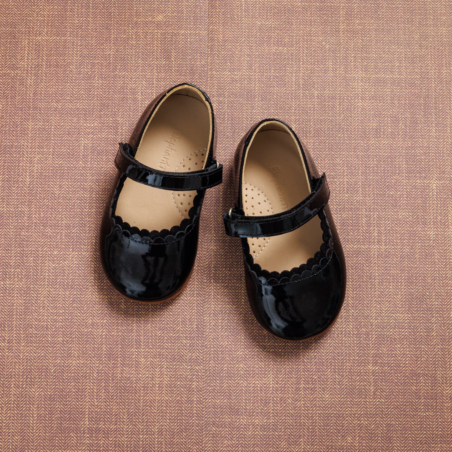 Black patent leather toddler Mary Jane shoes with scalloped edges and Velcro strap closure on textured brown background.