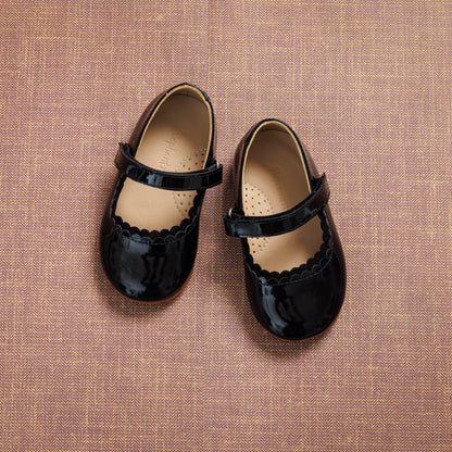Black patent leather toddler Mary Jane shoes with scalloped edges and Velcro strap closure on textured brown background.